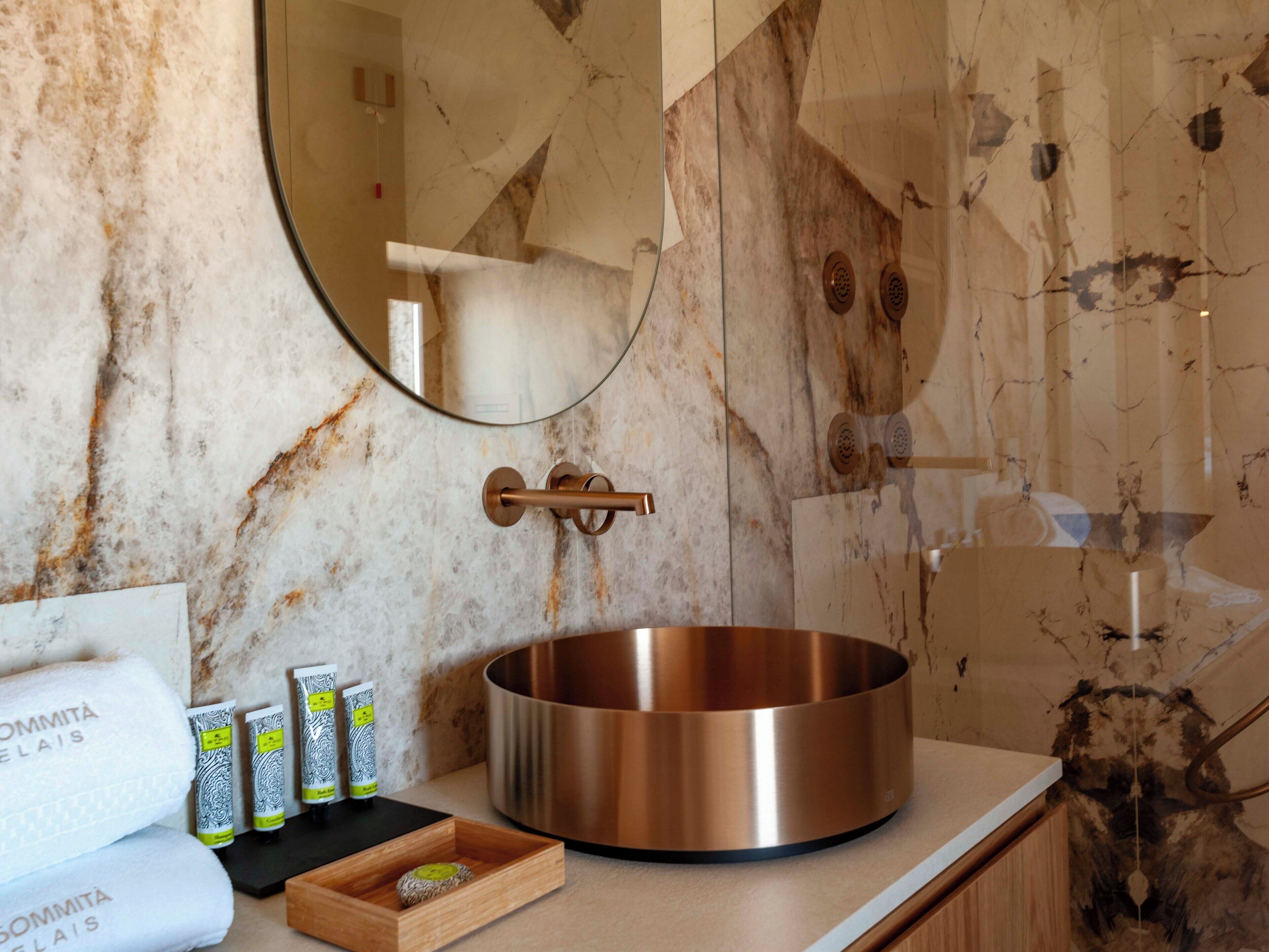 A bathroom with a marble wall, a round copper sink, a mirror, and toiletries on the counter.