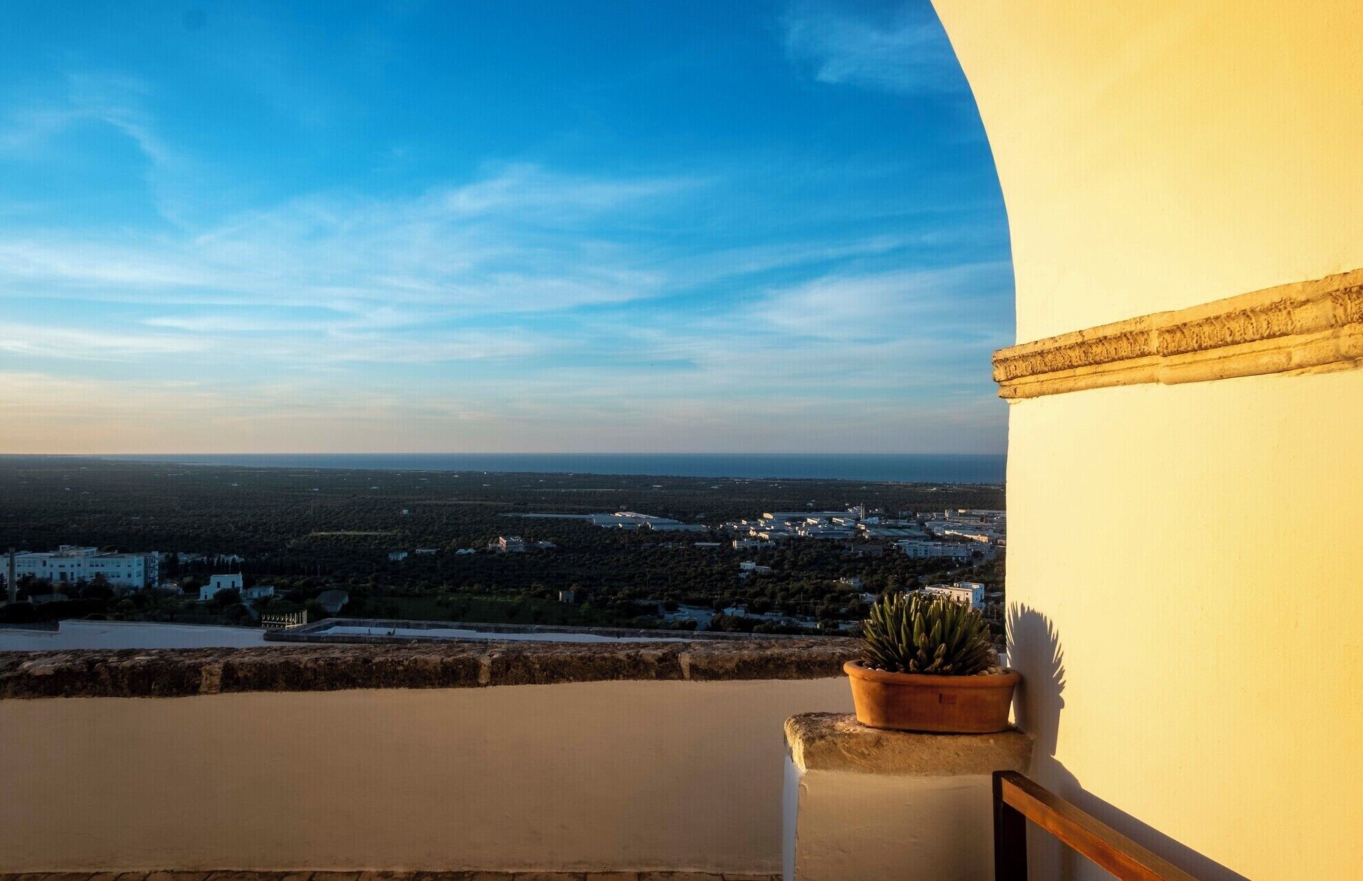 A balcony with a potted plant, overlooking a coastal landscape with buildings and a clear sky.