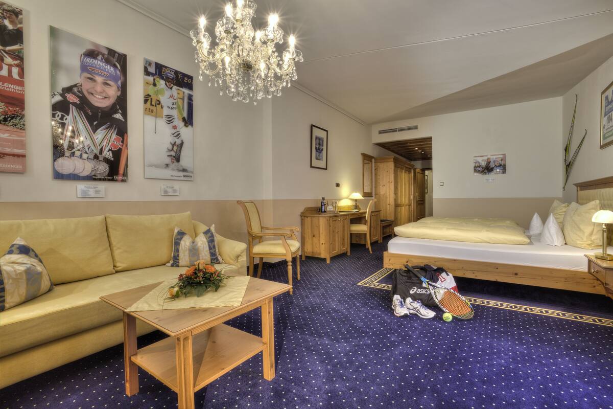 Ein Hotelzimmer mit Bett, Sofa, Schreibtisch, einer Chandelierleuchtre und Sportmemorabilien an den Wänden.