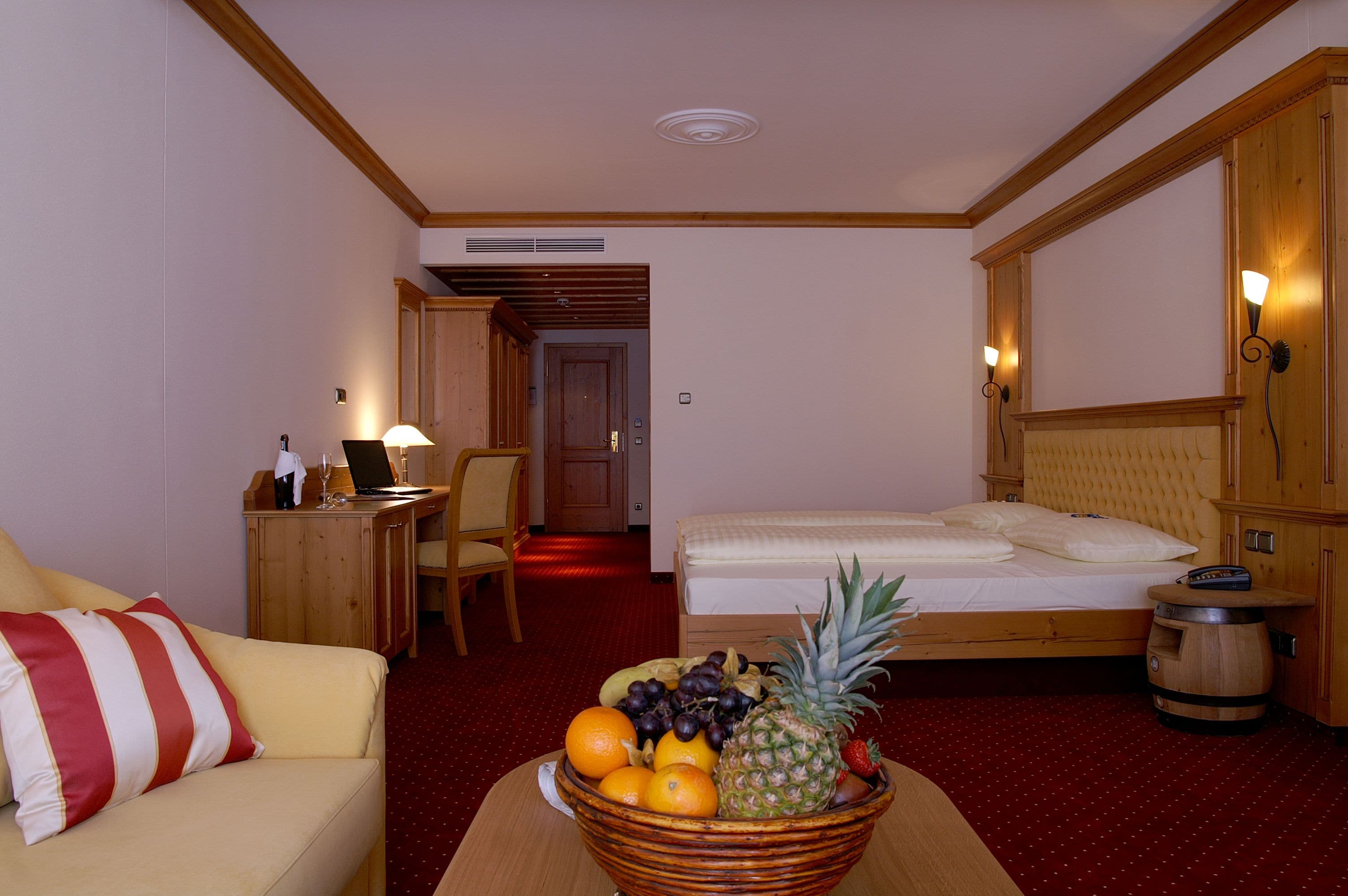 A hotel room with a bed, a wooden desk, a chair, a small table with a fruit bowl, and a red carpet.