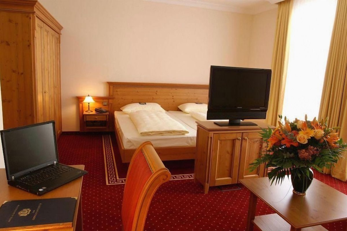 A hotel room with a bed, a desk with a laptop, a television, a nightstand with a lamp, and a vase of flowers.