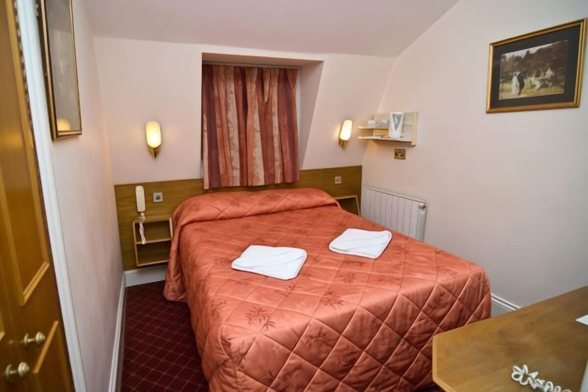 Habitación de hotel con cama, mesita de noche, ventana con cortinas y estante fijado a la pared.