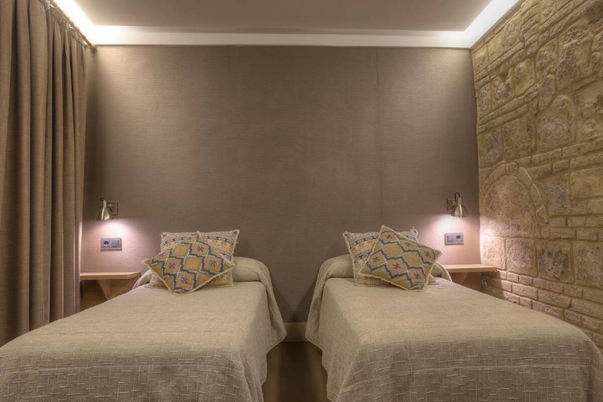 Ein Zimmer mit zwei Betten, jedes mit gemusterten Kissen, einer Steinsäule und einem Vorhang links.