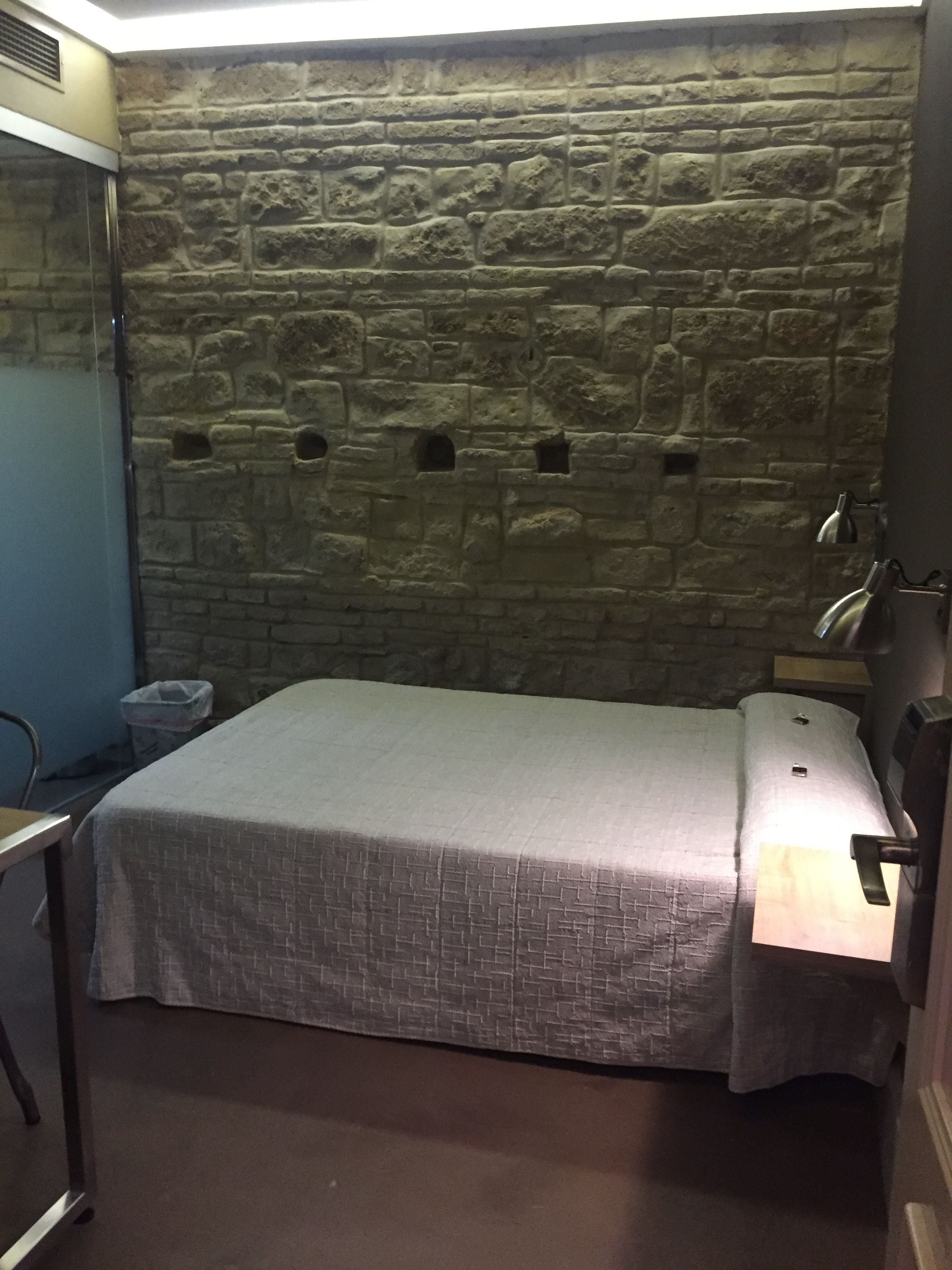 A room with a stone wall, a bed with a white bedspread, a bedside table with a lamp, and a chair.