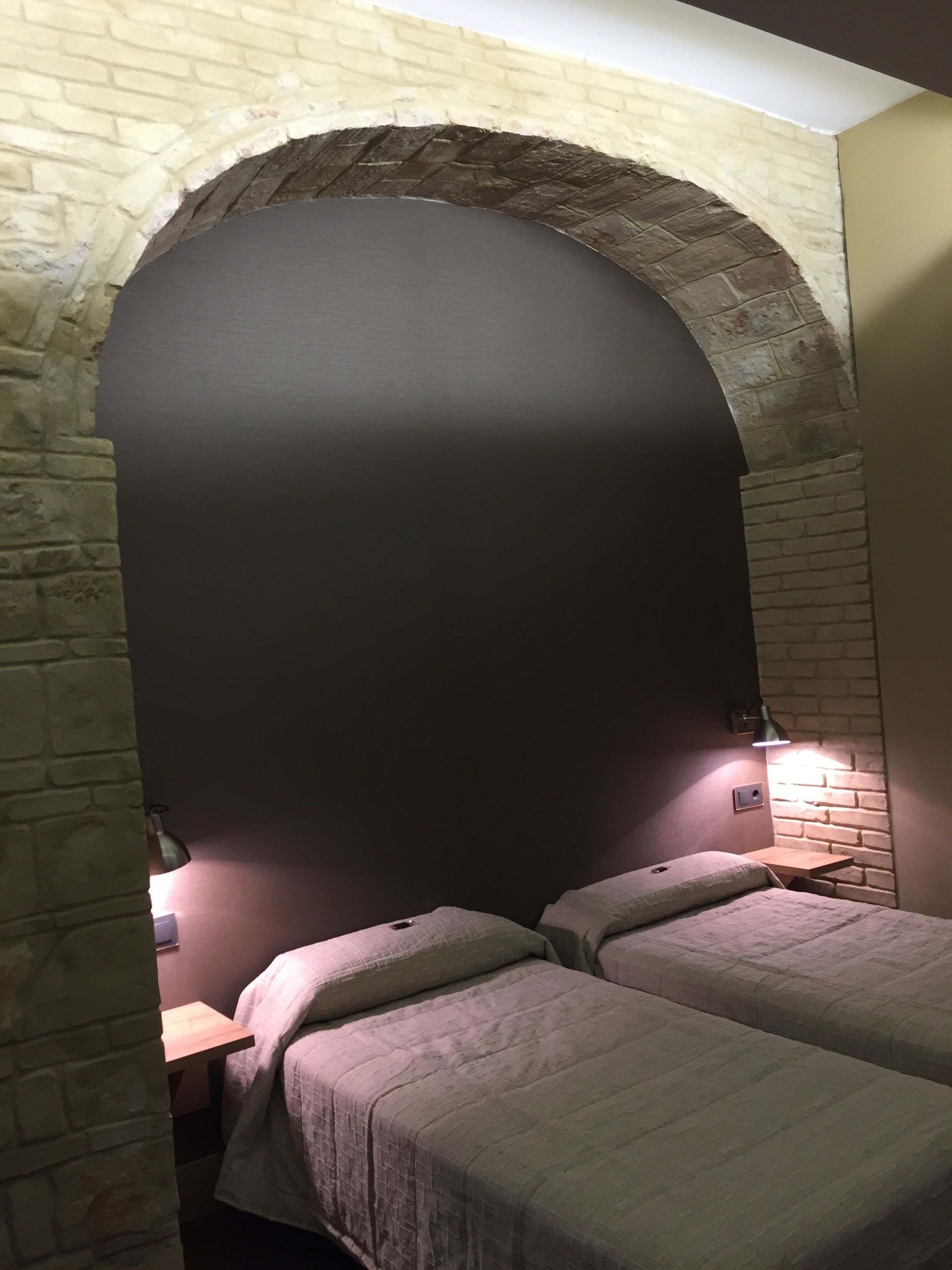 A room with a brick archway, two beds with headboards, a nightstand, and a wall-mounted lamp.