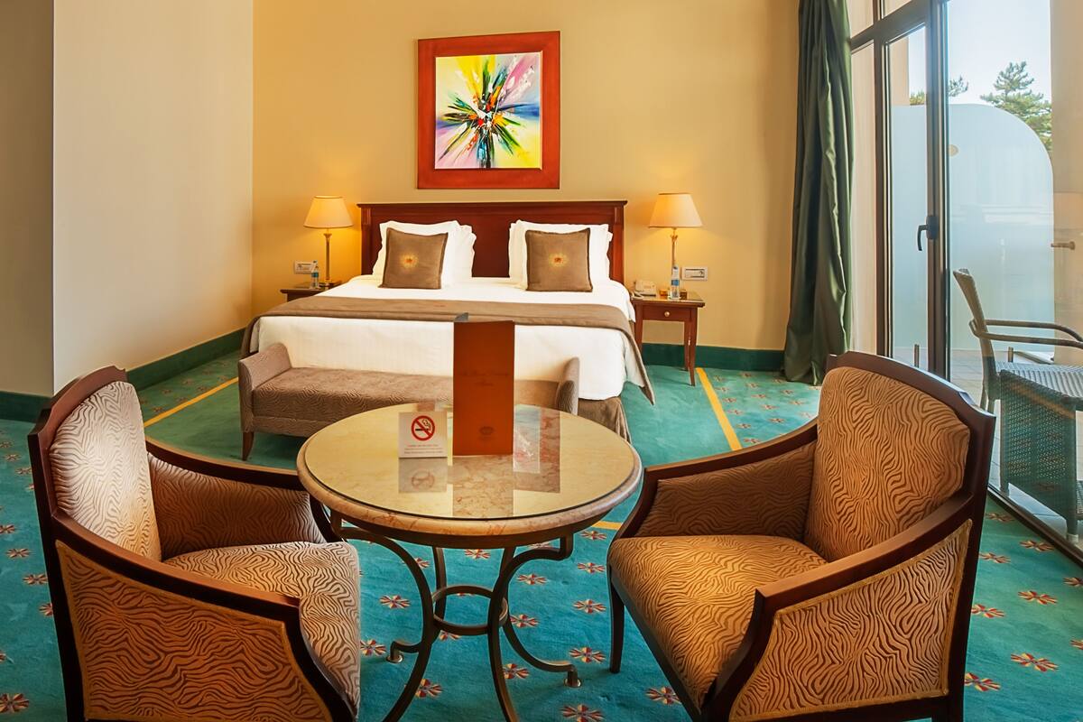 A hotel room with a bed, two bedside tables, a chair, a round table, and a large window.