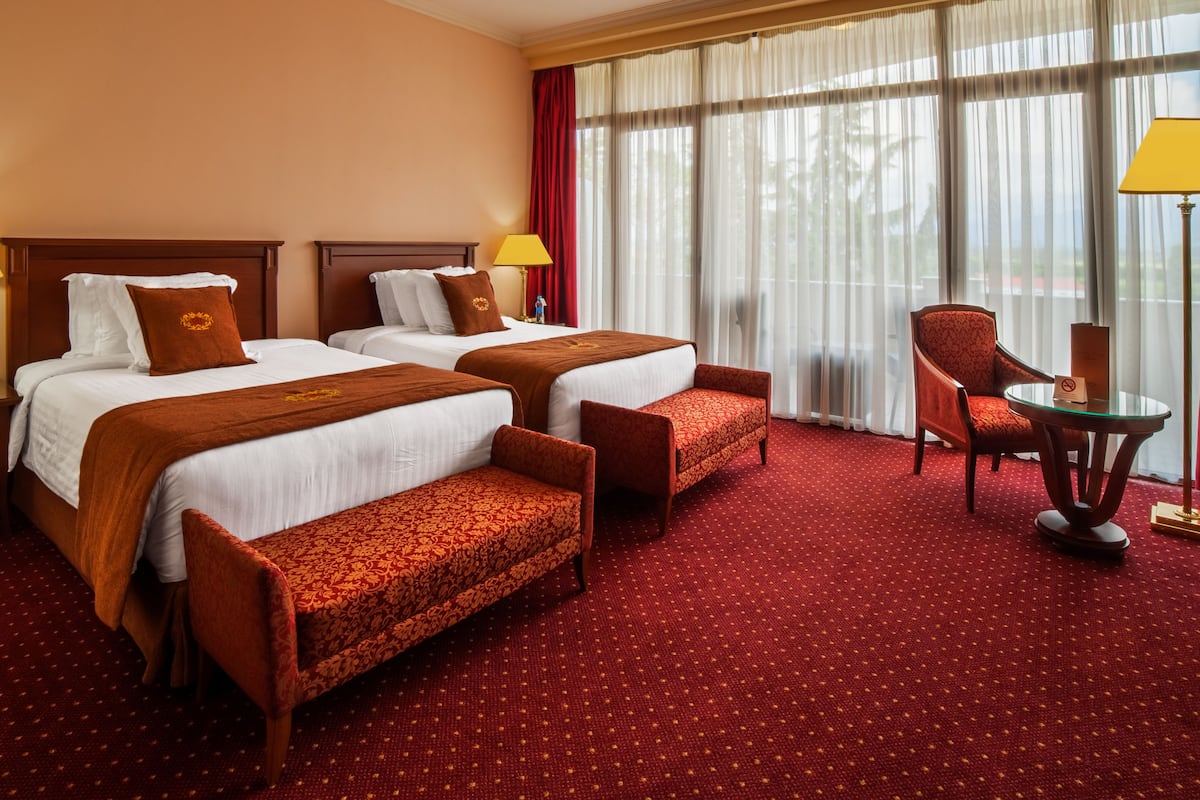 A hotel room with two beds, a sitting area, and a view of trees through sheer curtains.