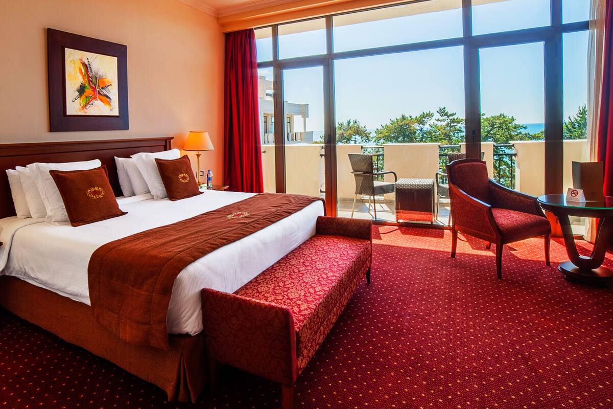 A hotel room with a large bed, two bedside lamps, a red carpet, a chair, a table, and a balcony with a view of the sea.
