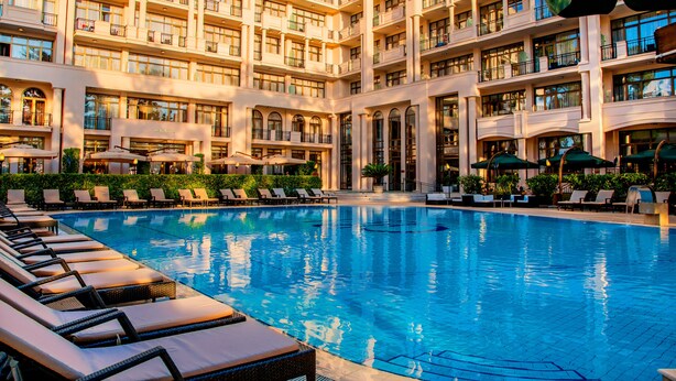 Une grande piscine intérieure, dotée de chaises longues et de parasols, et entourée d’un immeuble à plusieurs étages avec des balcons.