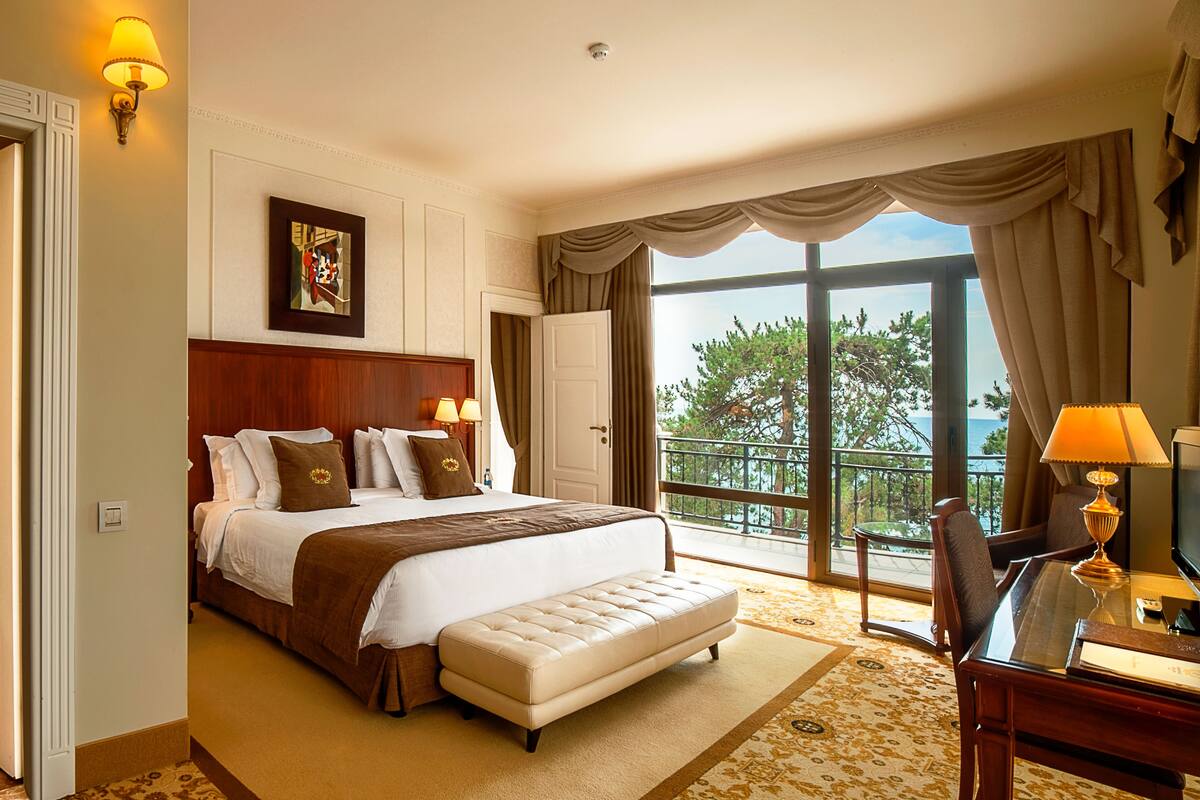 A hotel room with a large bed, a wooden headboard, a sitting area with a table and chair, and a balcony with a view of trees and the sea.