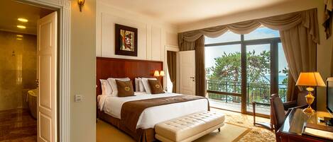 A hotel room with a large bed, a wooden headboard, a sitting area with a table and chair, and a balcony with a view of trees and the sea.