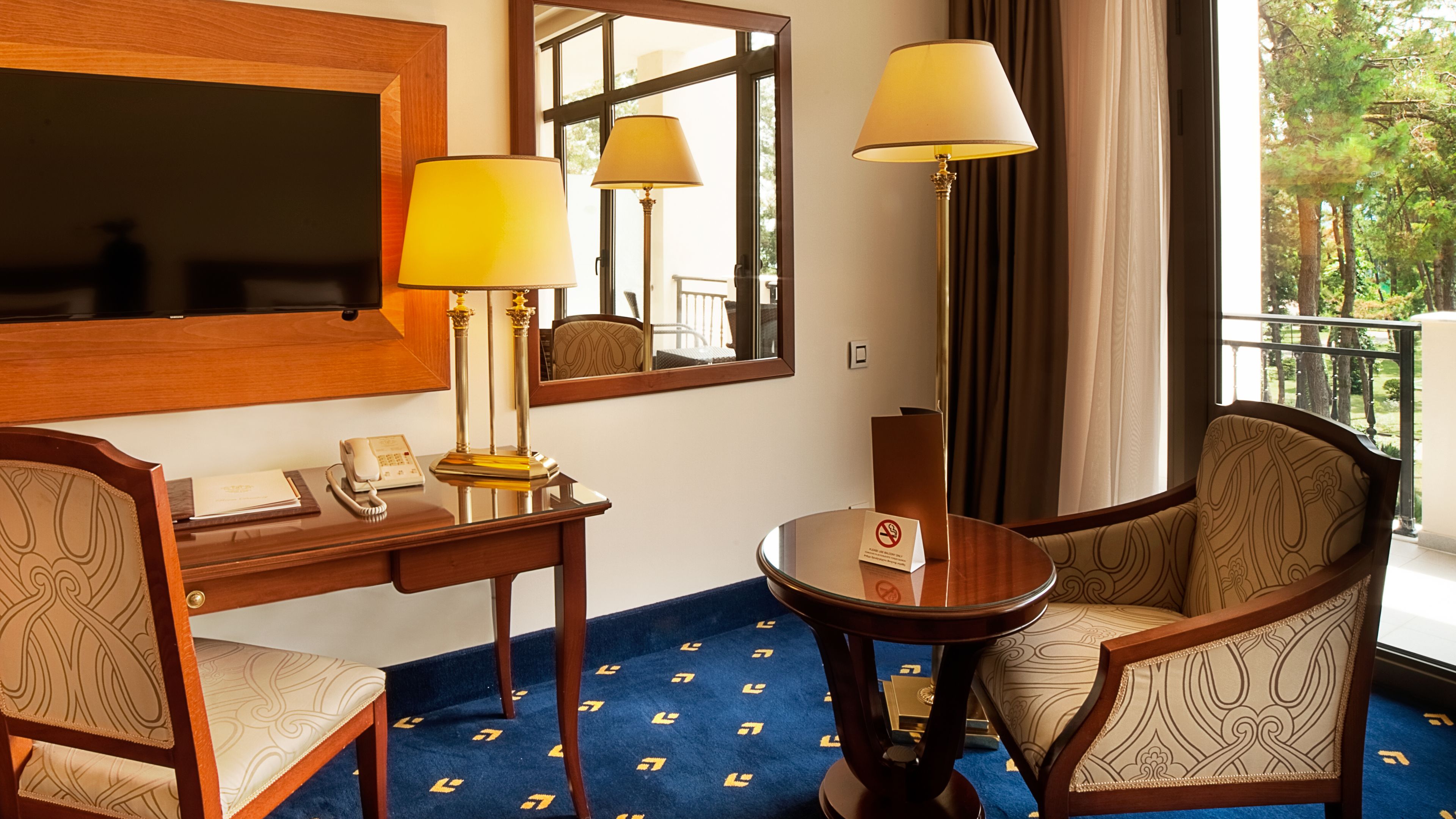 A hotel room with a desk, two lamps, a chair, a round table, and a view of the outdoors.