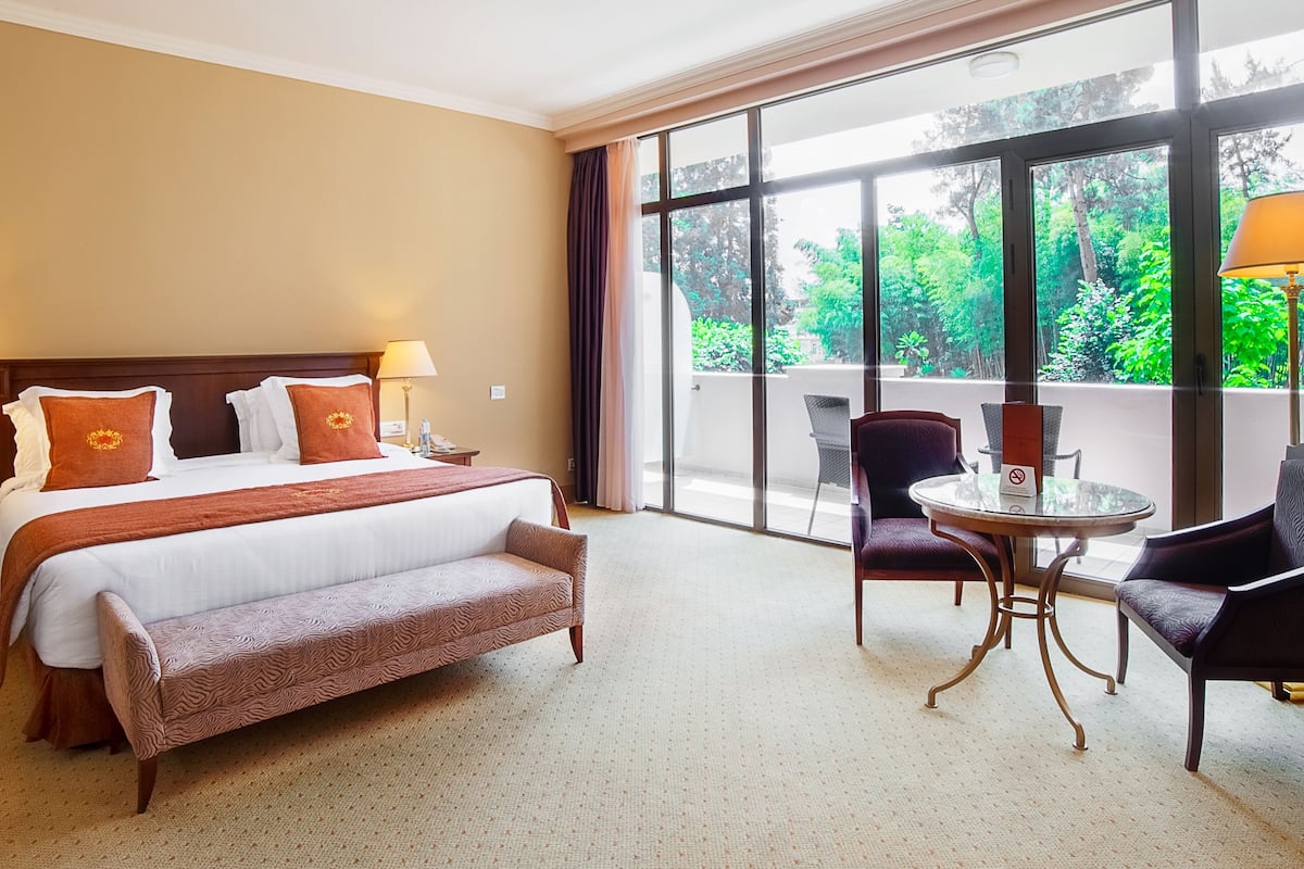 A hotel room with a large bed, two bedside lamps, a bench, a small table with chairs, and a balcony with a view of greenery.