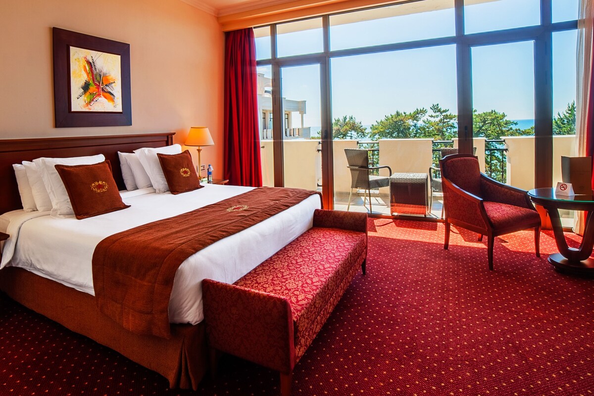Habitación de hotel con una cama grande, dos mesitas de noche, una silla, una mesa y un balcón con vistas.