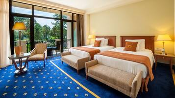 A hotel room with two beds, a sitting area with a chair and table, and a balcony with a view of trees.