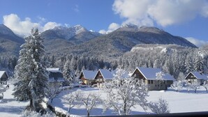 A snow-covered village with wooden houses, a prominent pine tree, and mountains in the background.