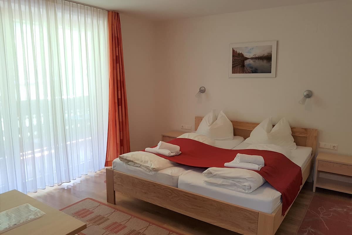 A neatly made bed with white linens and a red blanket, a wooden nightstand with a lamp, and a painting on the wall.
