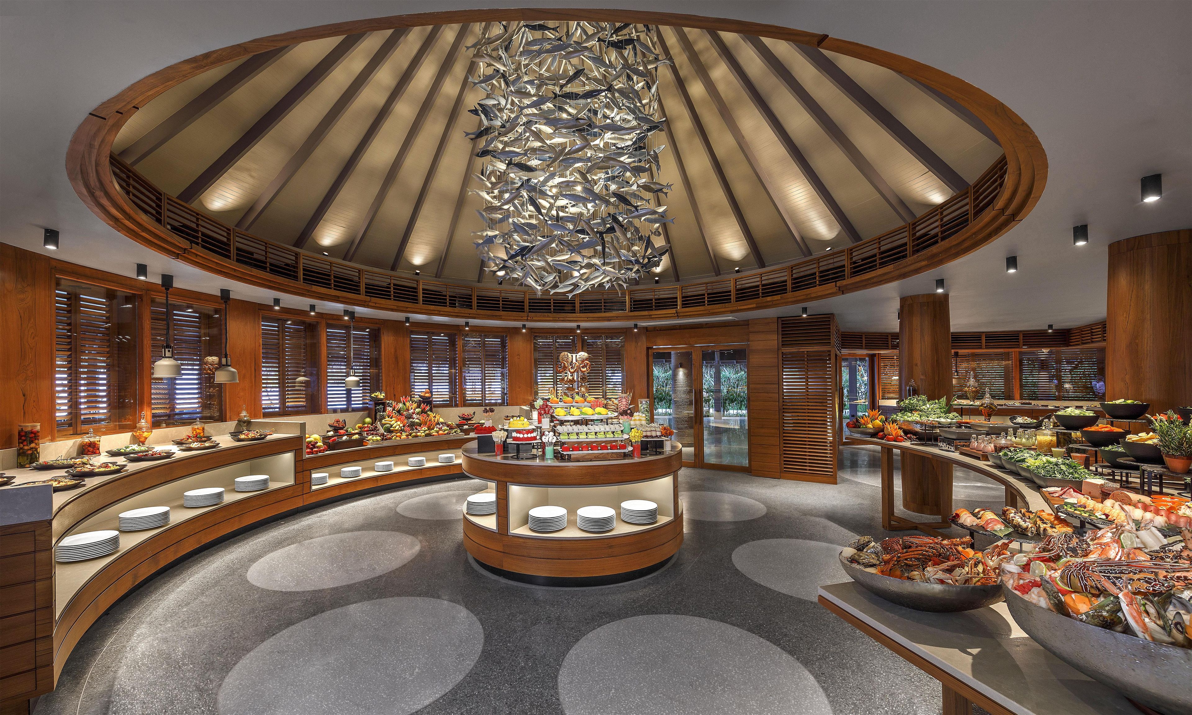A spacious hotel lobby with a central island featuring a variety of food and drink options, surrounded by wooden paneling and large windows.