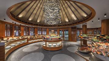 A spacious hotel lobby with a central island featuring a variety of food and drink options, surrounded by wooden paneling and large windows.