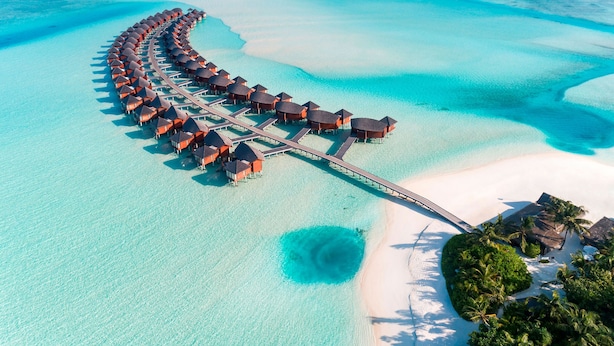 An aerial view of overwater bungalows connected by a bridge, surrounded by clear turquoise waters and white sand beaches.