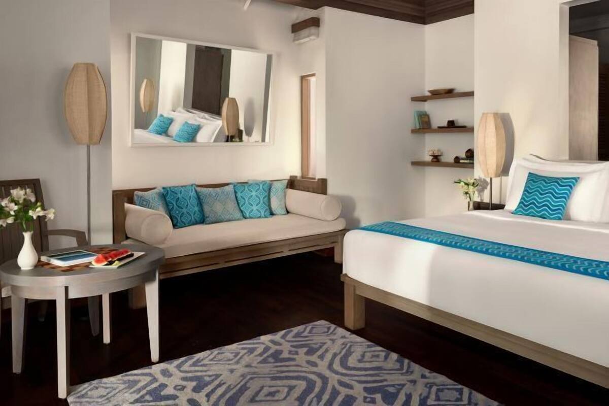 A hotel room with a bed, a sofa, a small table, a mirror, and a wall-mounted shelf.