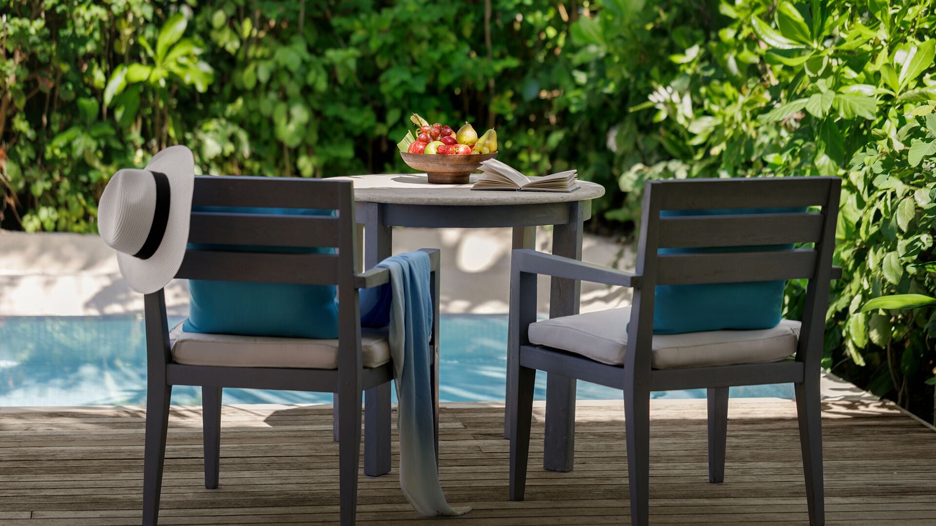 A wooden deck with two chairs and a table, a hat on one chair, and a bowl of fruit on the table.
