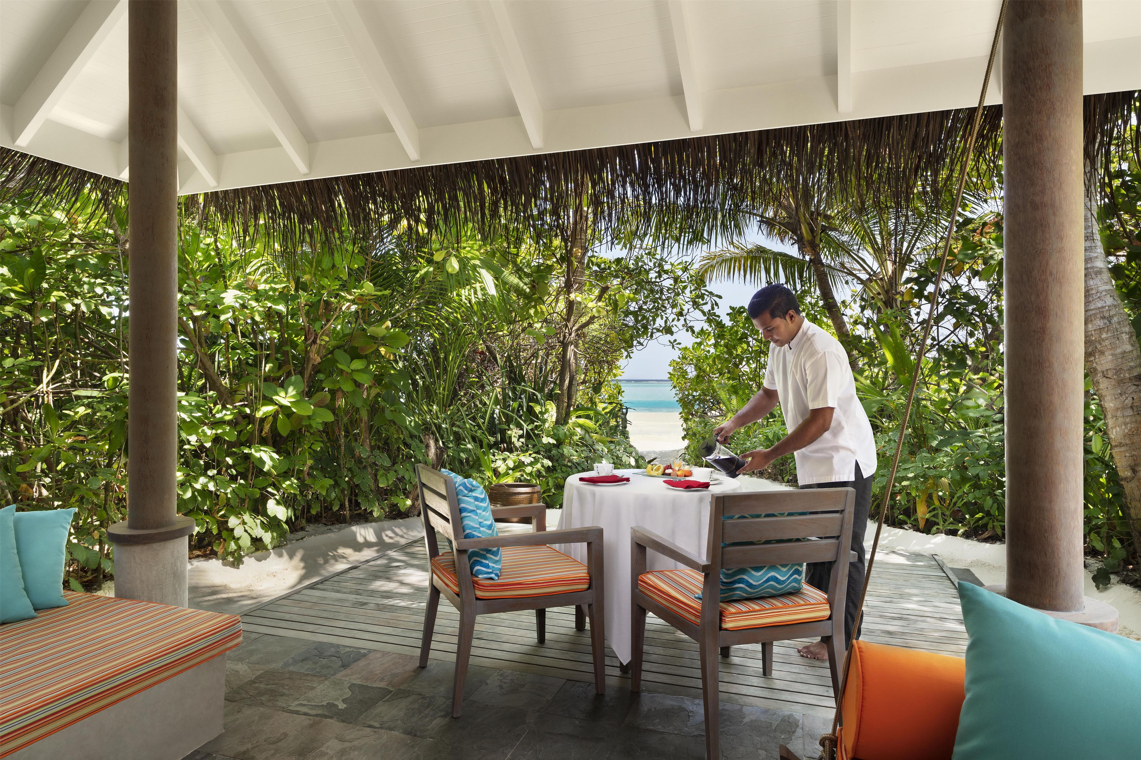 Un área de comedor al aire libre techada, con una mesa dispuesta para dos personas, rodeada de exuberante follaje y vista al mar.