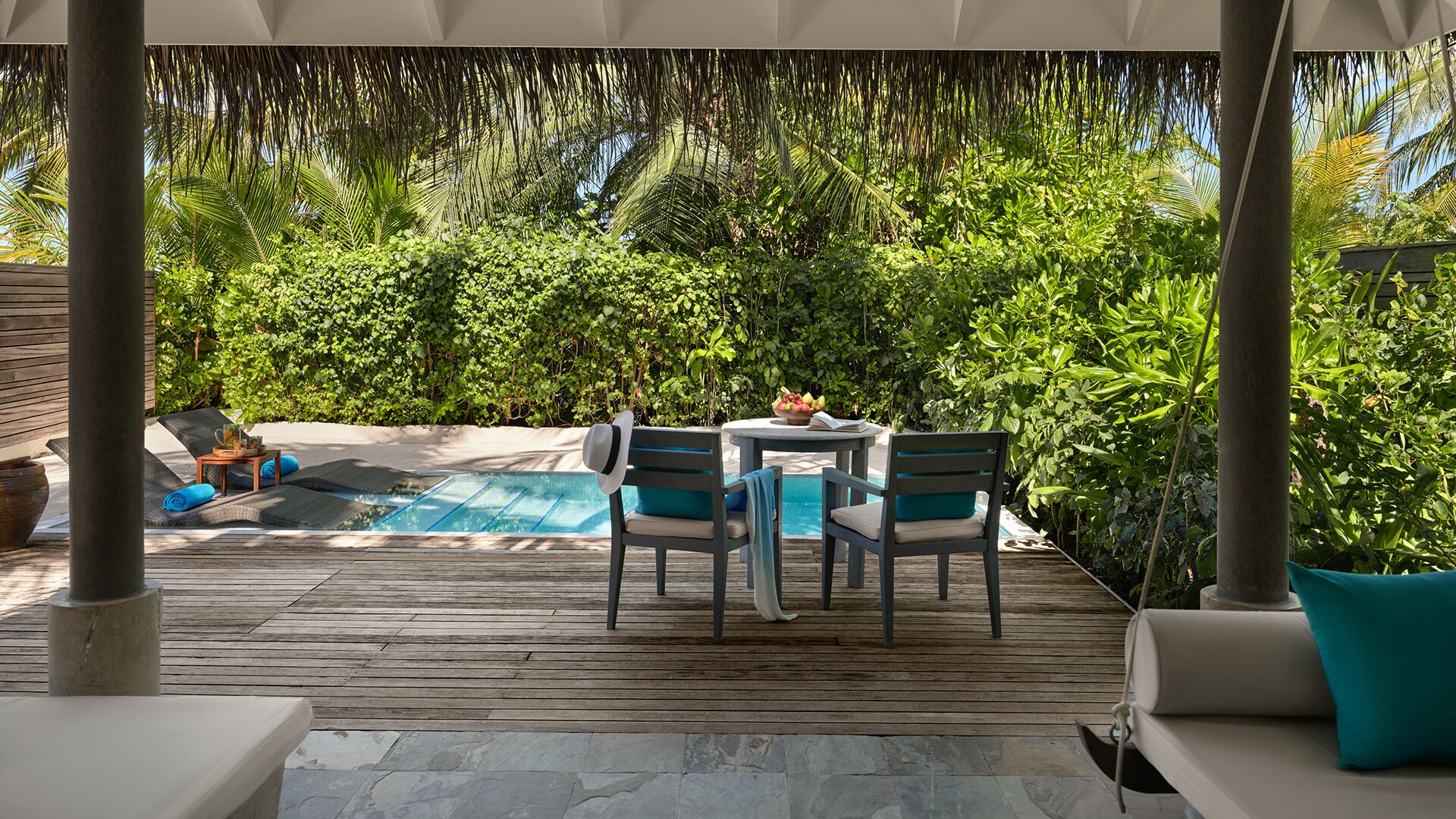 Um deck de madeira com piscina, rodeado por vegetação exuberante e um telhado de palha.