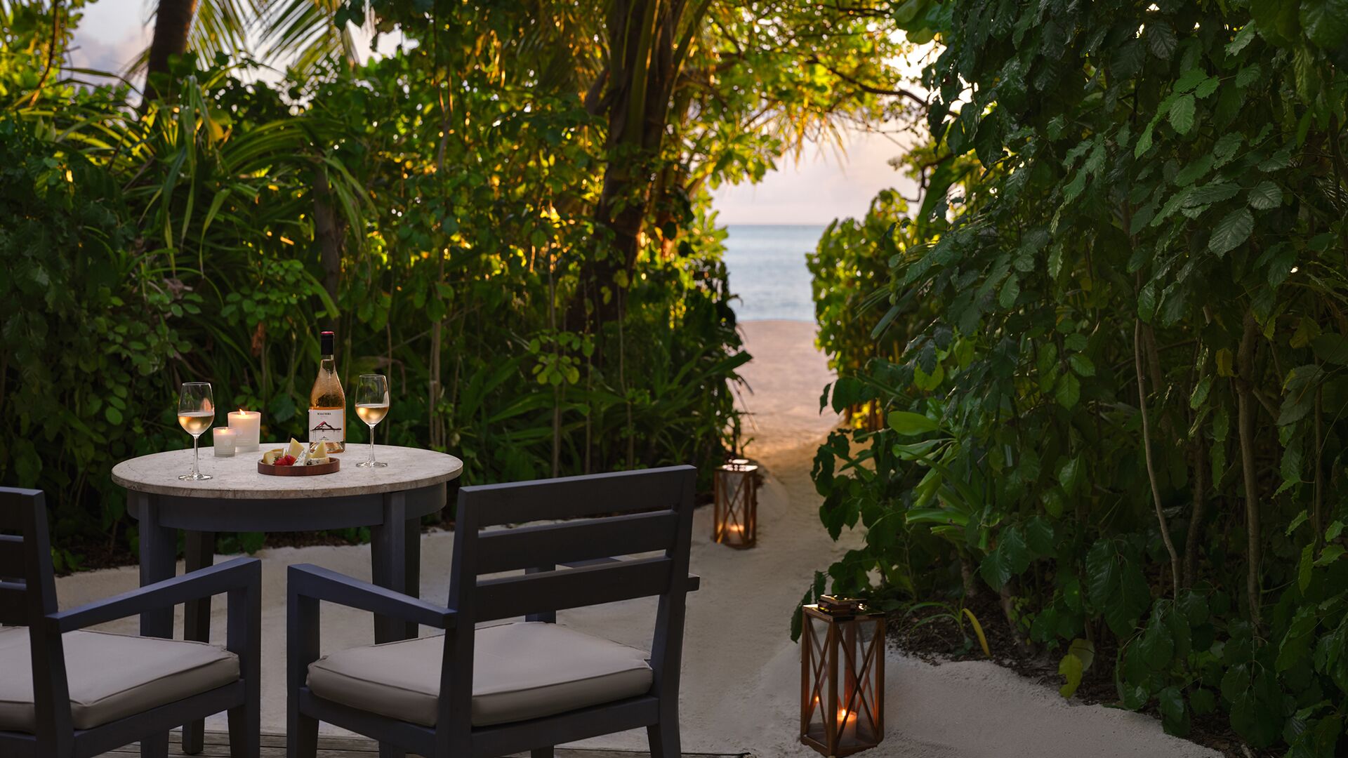 A table set for two with wine glasses and a bottle, surrounded by lush greenery and a view of the beach.