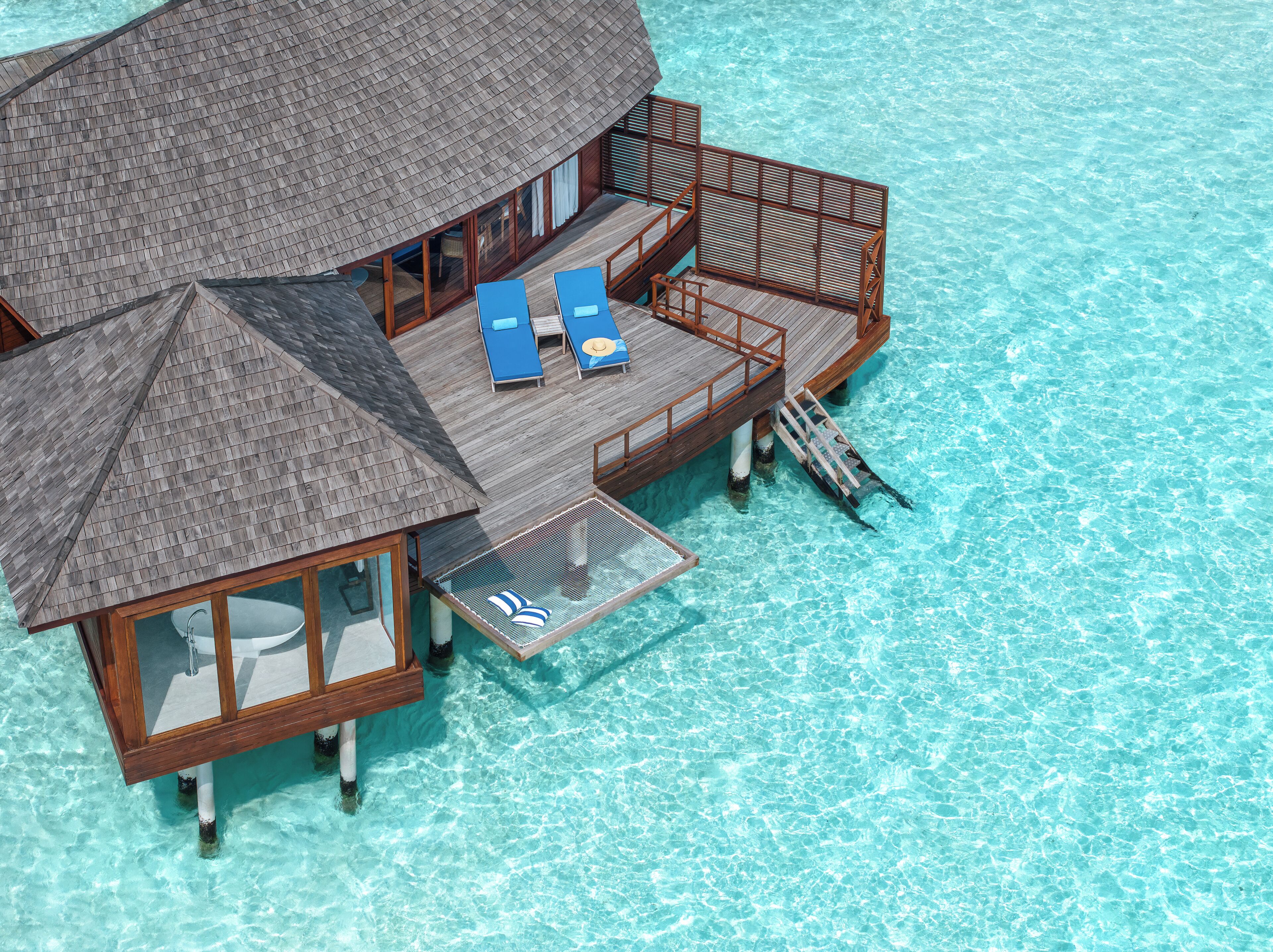 An overwater bungalow with a thatched roof, wooden deck, and two lounge chairs.