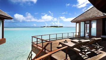 An overwater bungalows with wooden deck, lounge chairs, and a view of the turquoise sea and distant island.