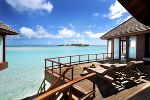 Bungalows sobre el agua con terraza de madera, sillas de descanso y vista al mar turquesa y una isla distante.