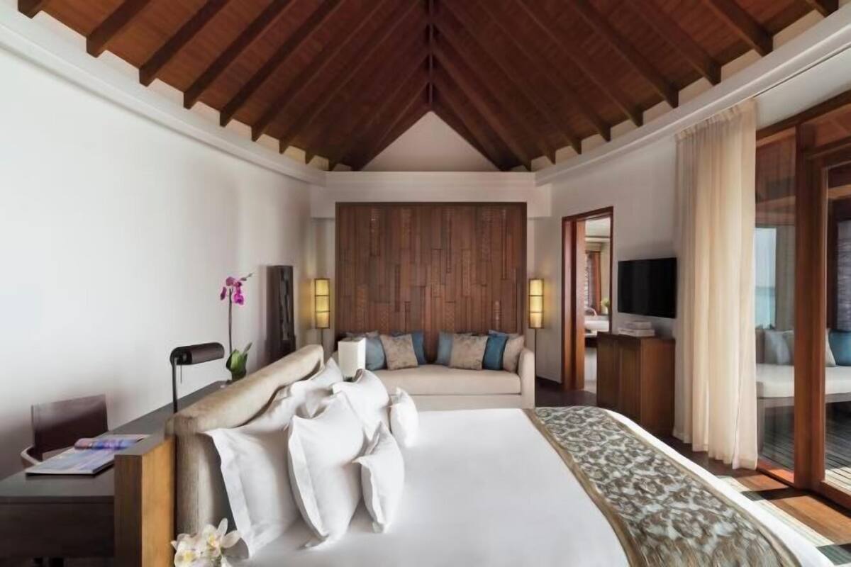 A hotel room with a wooden ceiling, a bed with white linens, a sofa, a TV, and a desk with a lamp.