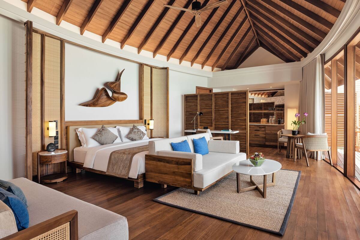 A spacious living area with a high ceiling, wooden beams, a large bed with white bedding, a sofa with blue cushions, a round coffee table, and a view of the outside through large windows.