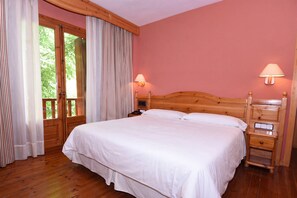 Un dormitorio con cama de madera, dos mesitas de noche, una mesita auxiliar, una ventana con cortinas y vista a zonas verdes.
