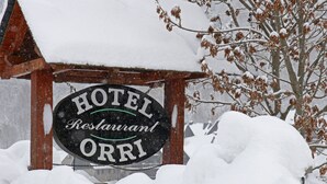 Señal de un restaurante de hotel llamado 'HOTEL ORRI', con estructura de madera rústica y acumulación de nieve alrededor.