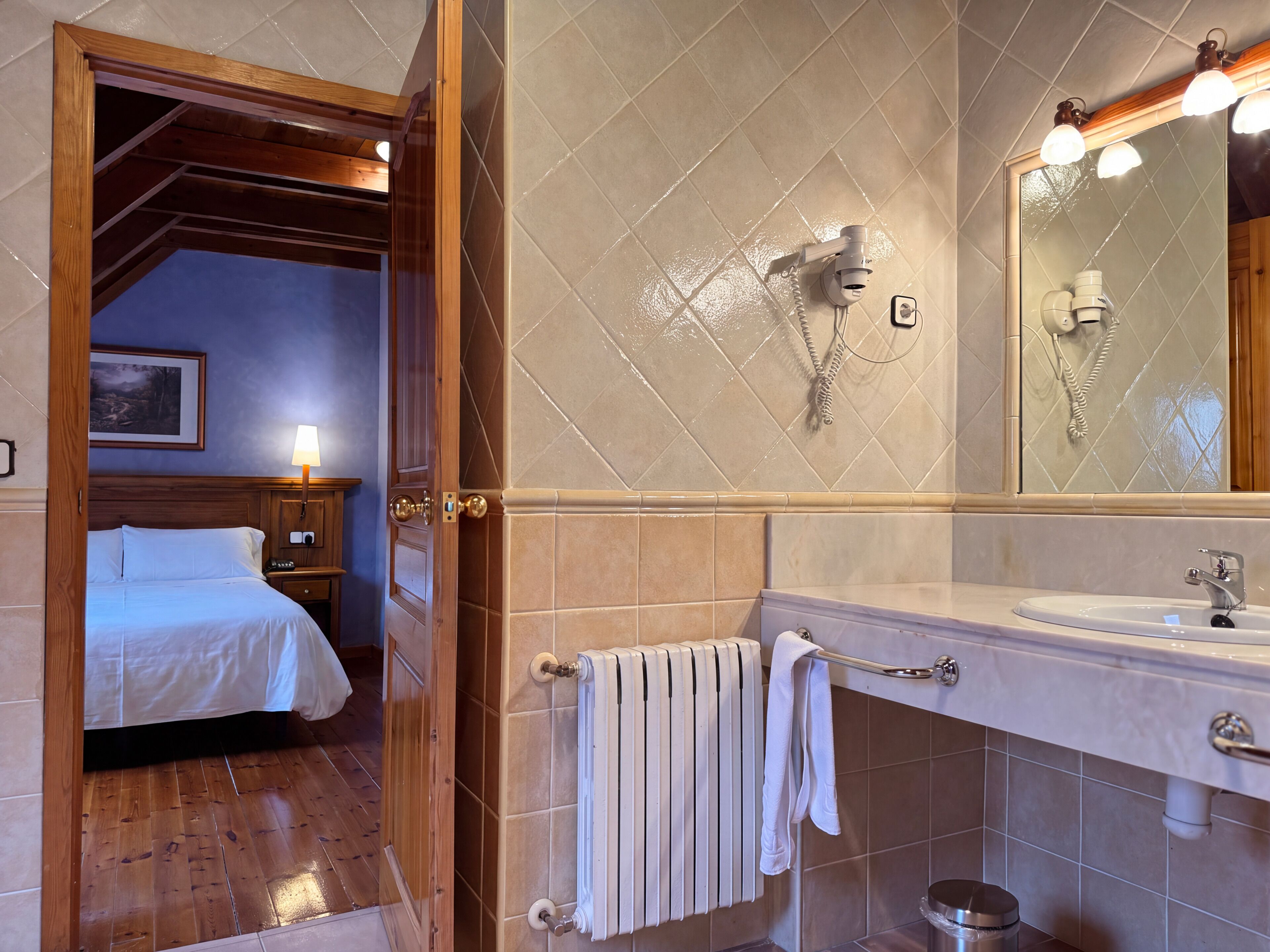 A hotel room with a wooden ceiling, a bed, a nightstand, a bathroom with a sink, a mirror, and a towel rack.