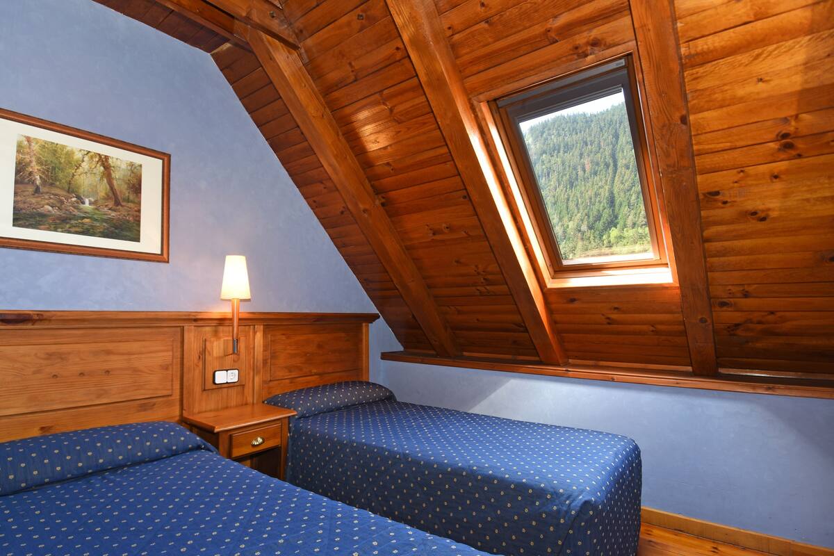 Una habitación con dos camas, cabecera de madera, una mesita de noche, una lámpara y una ventana con vista al bosque.