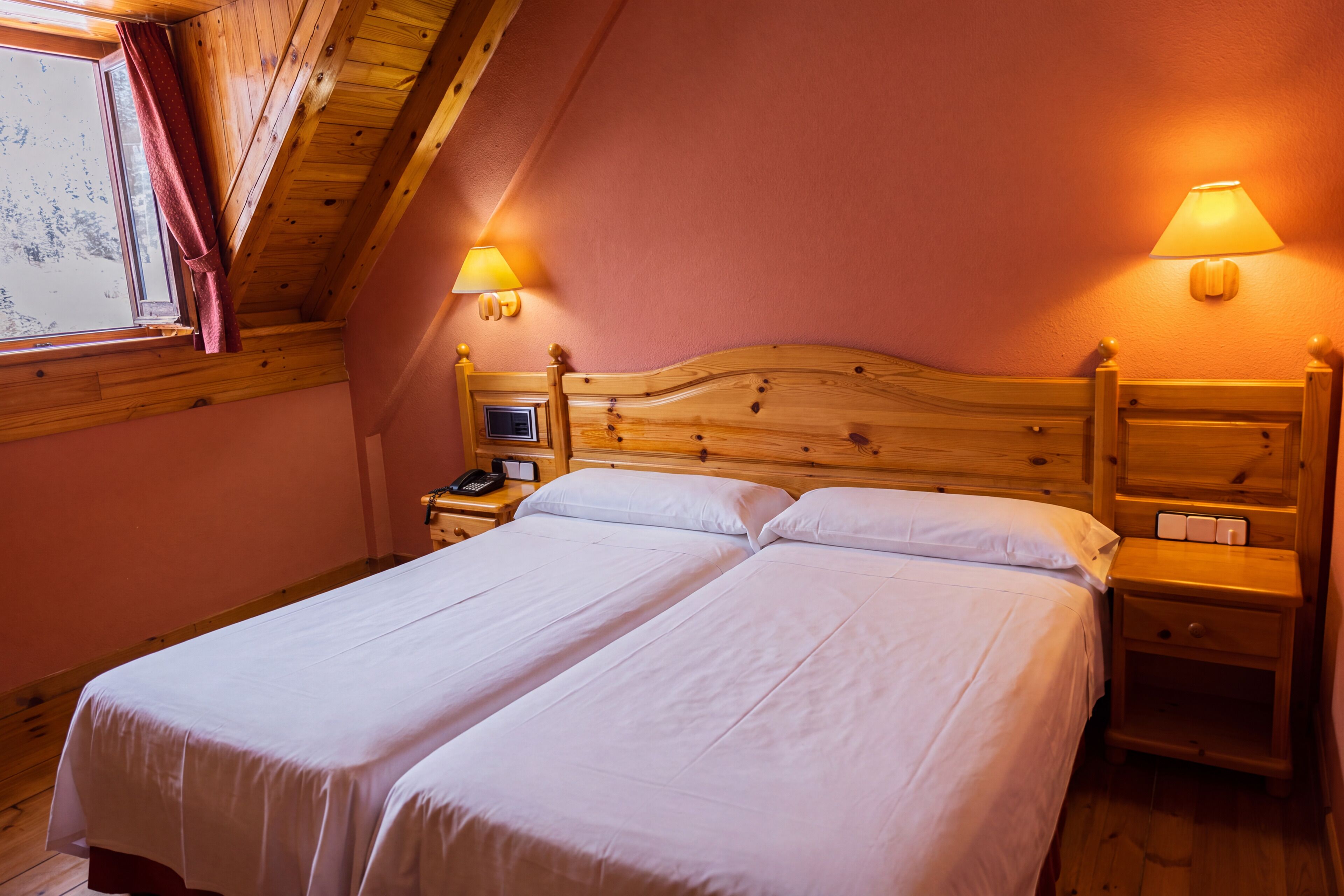 A double bed with two bedside tables, each with a lamp, in a room with wooden ceiling beams and a window with curtains.