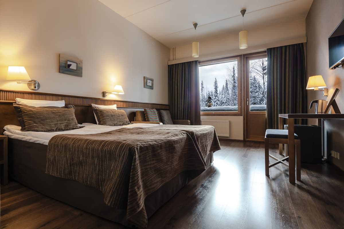 Habitación de hotel con una cama grande, lámparas de noche, un escritorio y vistas a árboles cubiertos de nieve a través de la ventana.