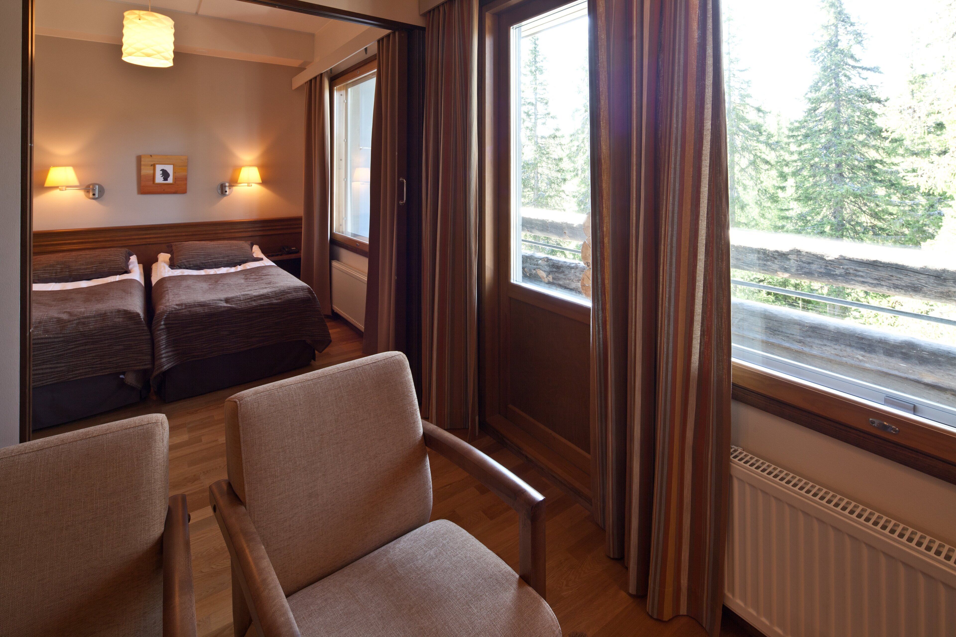 Habitación de hotel con dos camas, una silla, una ventana con cortinas y vista a un bosque.