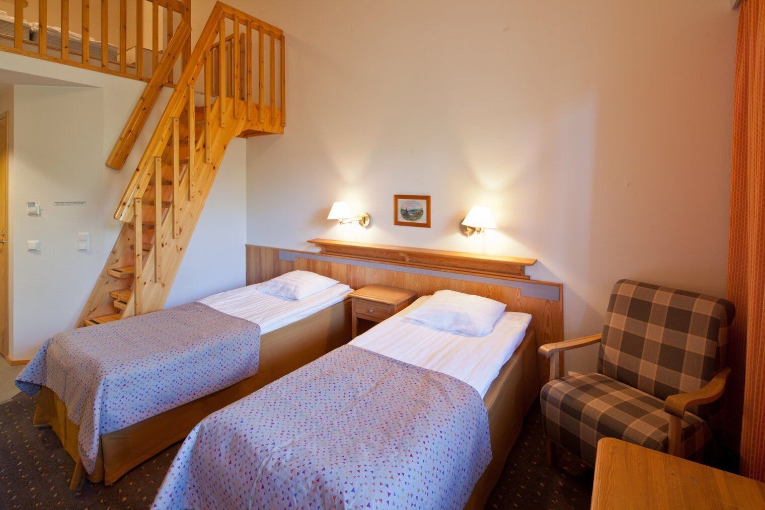 Una habitación con dos camas individuales, una escalera de madera, un sillón a cuadros y una mesita de noche con una lámpara.