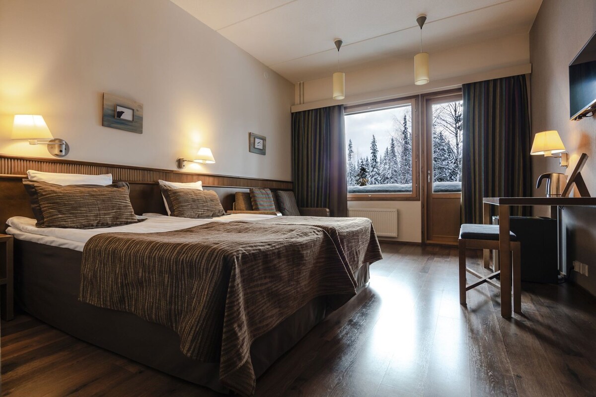 A hotel room with a large bed, bedside lamps, a desk, and a view of trees through the window.