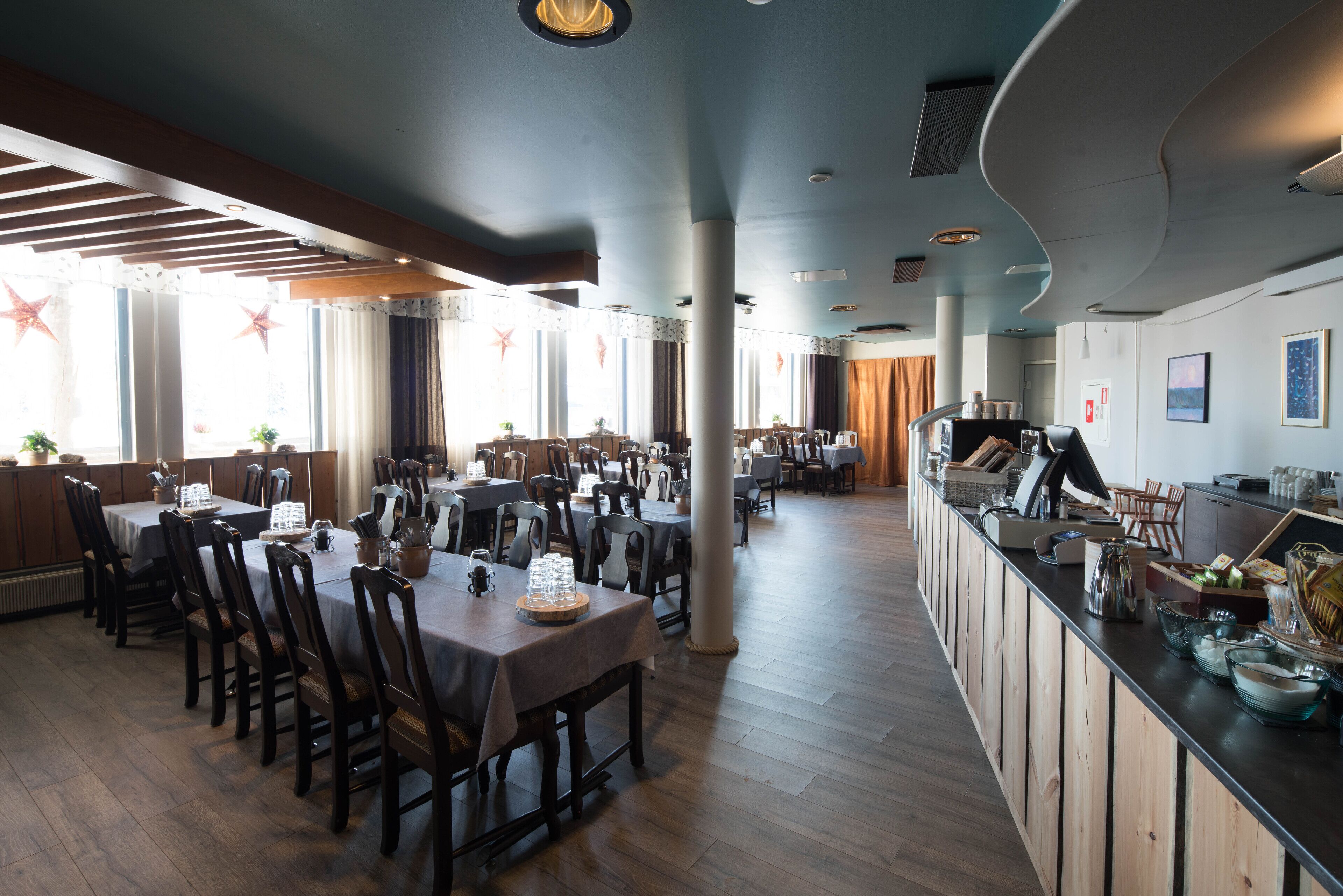 Un restaurante con mesas y sillas de madera, una zona de bar y amplias ventanas con cortinas.