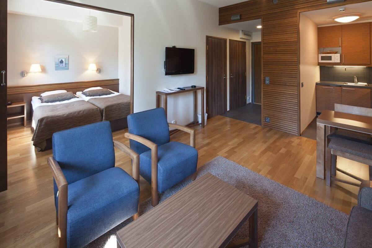 Habitación de hotel con una cama, dos sillones azules, una mesa de madera, televisor, una mesita con papeles, microondas y zona de cocina.