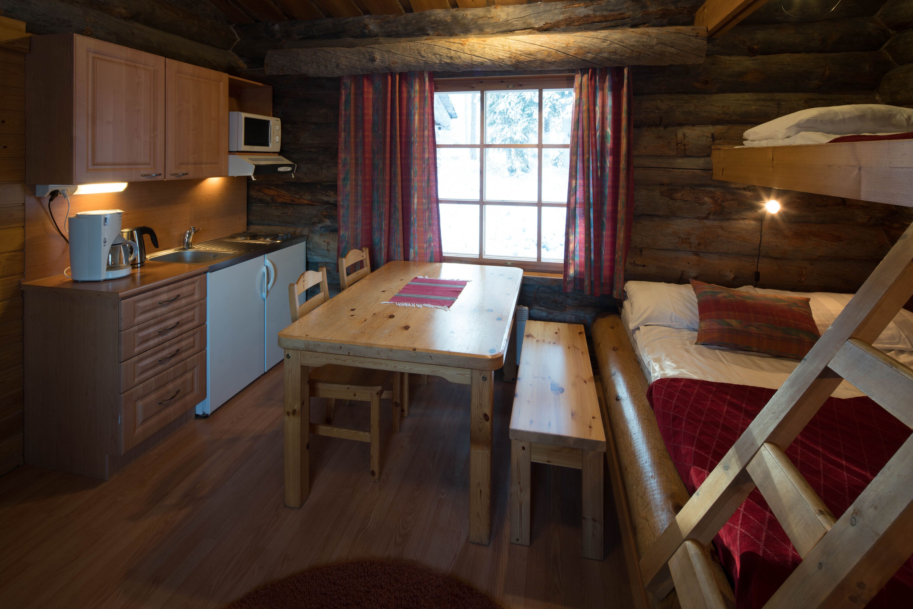Une cabane chaleureuse avec un intérieur en bois, une table à manger, des bancs, un lit superposé et un coin cuisine équipé d’un four à micro-ondes.