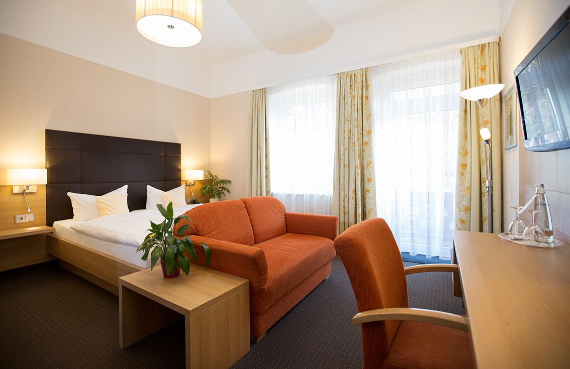 Ein Hotelzimmer mit einem Bett, einem Sofa, einem Schreibtisch und einem Sessel. Auf den Nachttischen stehen zwei Lampen, auf dem Sofa ein Blumentopf und an der Wand ein Fernseher.