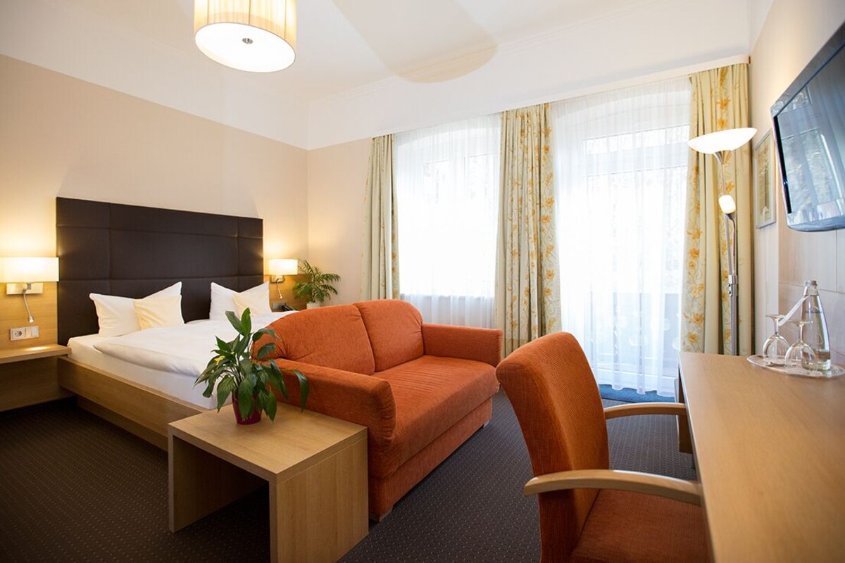 A hotel room with a bed, a sofa, a desk, and a chair. There are two lamps on the nightstands, a potted plant on the sofa, and a TV on the wall.