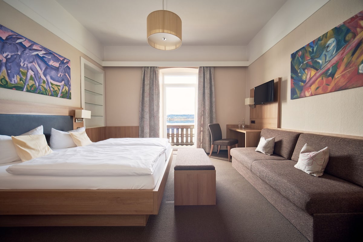 Quarto de hotel com uma cama grande, um sofá, uma mesa e uma televisão. Há quadros nas paredes e uma janela com vista.