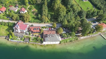 Vue aérienne d’une propriété au bord du lac, comprenant plusieurs bâtiments, dont un grand bâtiment blanc avec un toit rouge, le tout entouré d’une forêt dense et bordé d’un lac aux eaux claires.